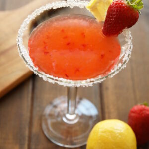A martini glass full of strawberry lemon drop martini sits on a wooden table. The glass has a sugared rim and is garnished with a wedge of lemon and a strawberry. The table has an additional lemon and strawberry on it.