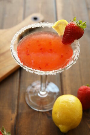 A martini glass full of strawberry lemon drop martini sits on a wooden table. The glass has a sugared rim and is garnished with a wedge of lemon and a strawberry. The table has an additional lemon and strawberry on it.
