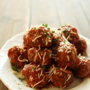 A big white plate is resting on an old fashioned wooden table. The plate is piled high with homemade Italian meatballs that are sprinkled with Parmesan cheese and fresh herbs.