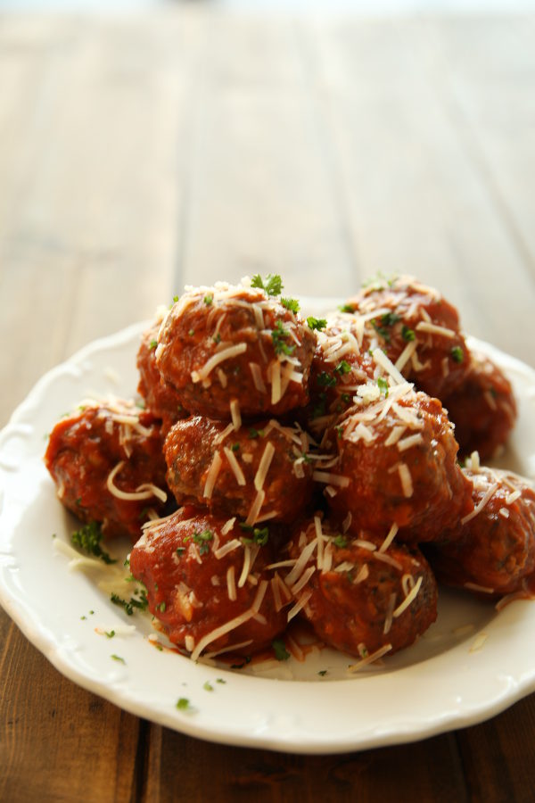 A big white plate is resting on an old fashioned wooden table. The plate is piled high with homemade Italian meatballs that are sprinkled with Parmesan cheese and fresh herbs.