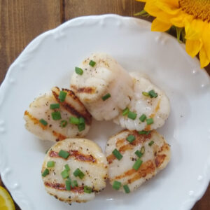 A white plate has 5 sea scallops arranged on it. The scallops are grilled and garnished with chive butter. There are some squeezed lemon wedges in one corner and a sunflower in another.