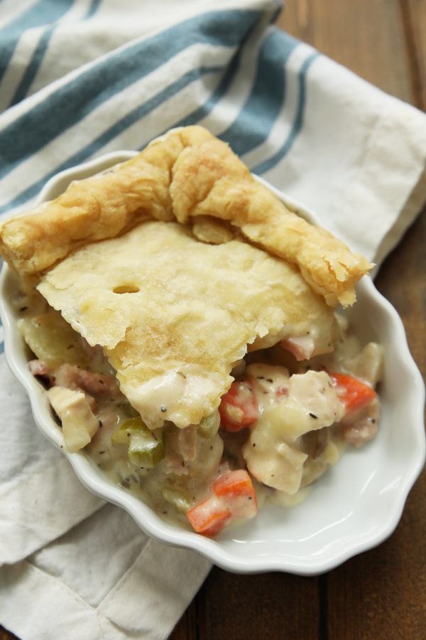 a slice of ham and chicken pot pie in a white dish. The dish is sitting on a white and blue stripped dish towel on a wooden table.