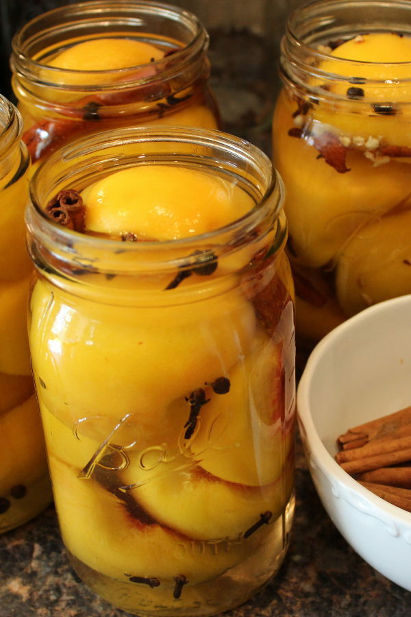 Mason jars sit on the counter next to a bowl of cinnamon sticks. The jars are being packed with peach halves and whole spices.