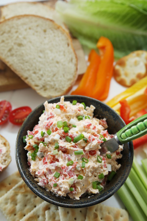 A pottery bowl of homemade pimento cheese is surrounded by saltines, celery, strips of bell pepper, crackers, lettuce, French bread, and sliced tomatoes.