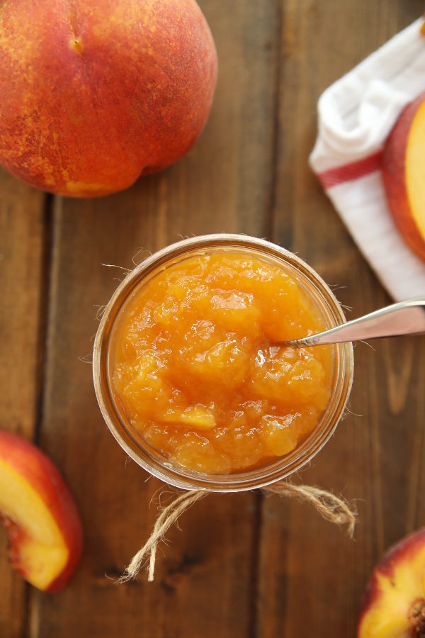 A mason jar of peach jam is open with a spoon sticking out. Nearby there are some fresh peaches sitting on a wooden table.