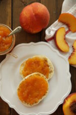 An English muffin is slathered with peach jam on a white plate. Nearby there is a mason jar of peach jam and some fresh peaches. j