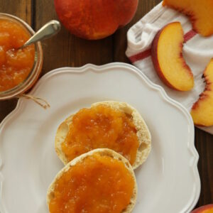 An English muffin is slathered with peach jam on a white plate. Nearby there is a mason jar of peach jam and some fresh peaches. j