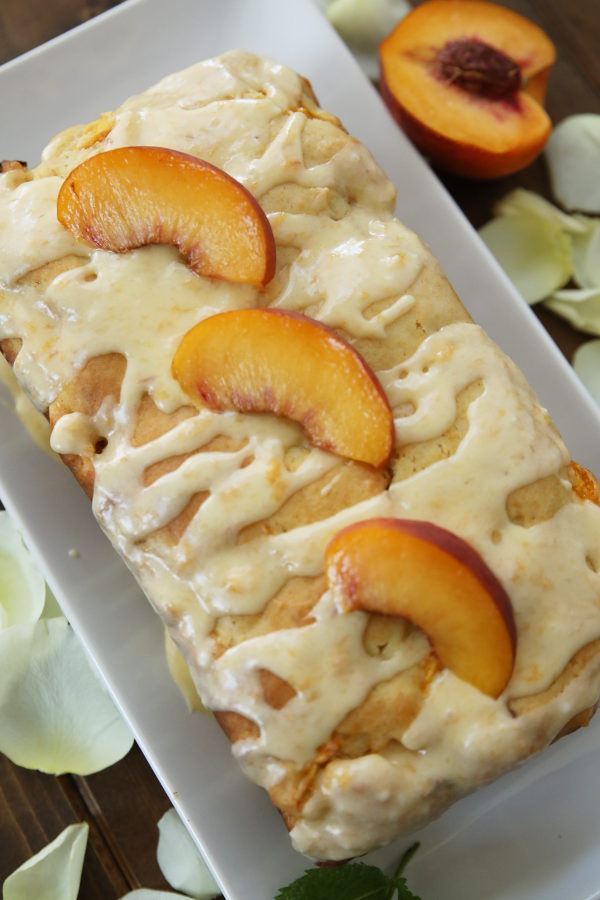 A loaf of fresh peach bread sits on a rectangular white plate. The bread is drizzled with a lot of fresh peach glaze which is a creamy yellow. Three slices of peach are on top of the bread. Next to the plate is a half a peach and some white rose petals scattered on the table.