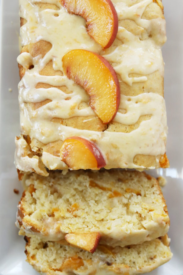 This photo is a close up of a loaf of peach bread. The first few slices have been cut and are resting at the end of the loaf. You can see the texture of the fresh peaches marbled into the bread. Slices of fresh peach sit on the glaze on top of the bread.