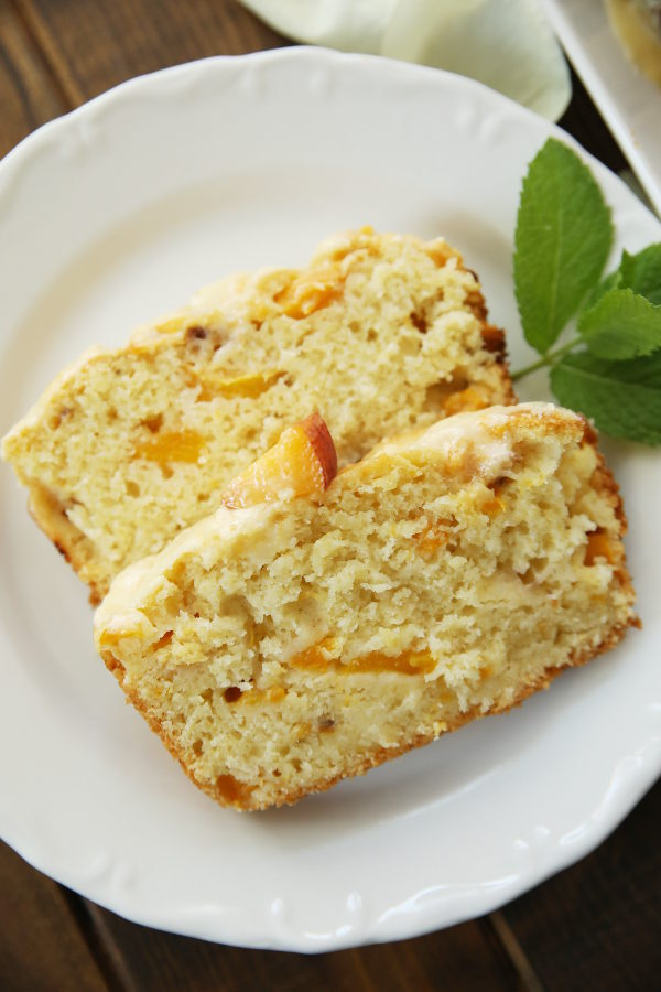 Two slices of peach bread sit on a white plate. A sprig of mint is decorating the plate. You can see the beautiful texture of the peach colored bread. The bread has a beautiful crumb studded with fresh peaches.