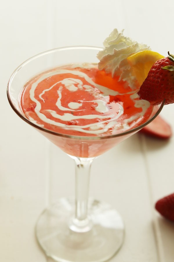 A vibrant, red strawberry martini fills a clear martini glass on a white table. The drink is garnished with heavy cream, a wedge of lemon and a strawberry.