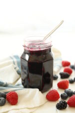 A clear mason jar is full of mixed berry jam. The jam is a dark purple and has a spoon in the top ready for serving. The jar of jam is on a table by a white and blue striped cloth and surrounded by raspberries, blueberries, and blackberries.