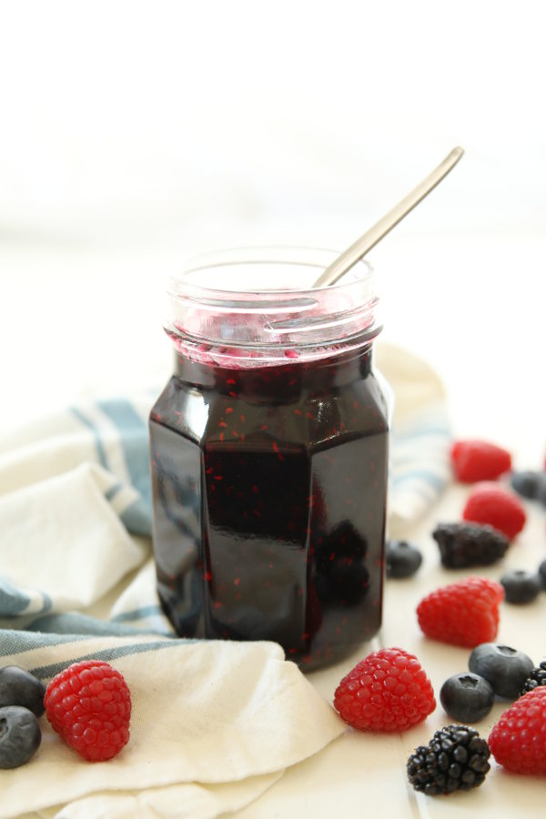 A clear mason jar is full of mixed berry jam. The jam is a dark purple and has a spoon in the top ready for serving. The jar of jam is on a table by a white and blue striped cloth and surrounded by raspberries, blueberries, and blackberries.