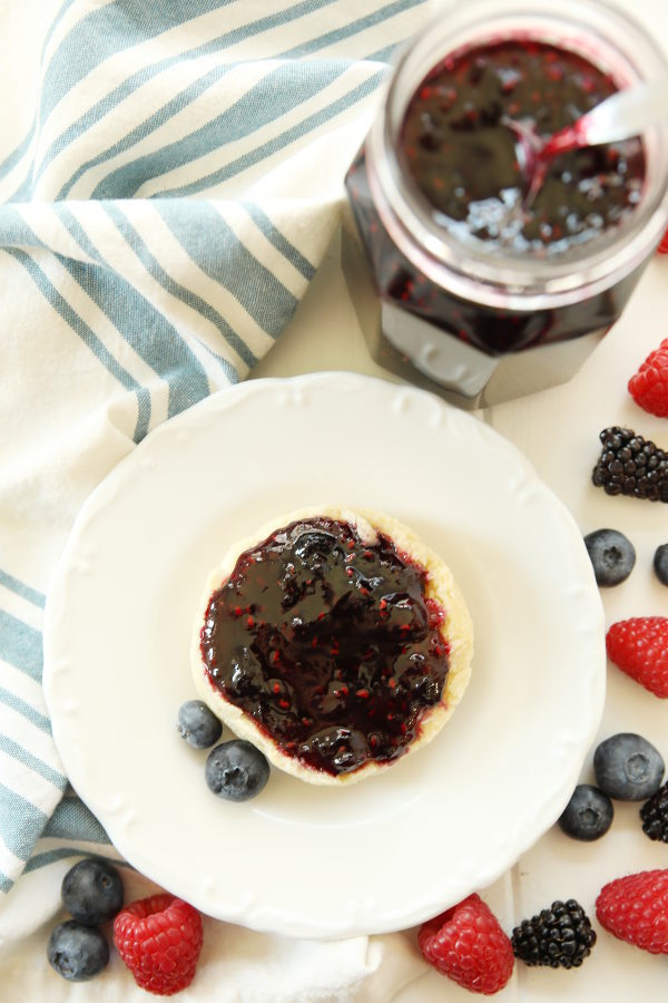 Half a biscuit is spread with mixed berry jam and sits on a white plate. Berries are scattered around the table and a mason jar of three berry jam sits next to the plate.