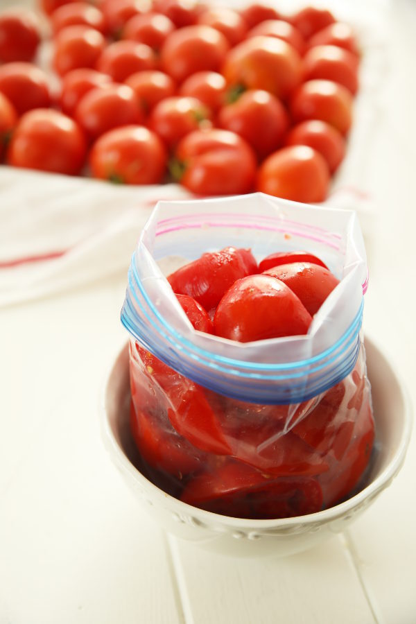 A quart ziploc bag is full of sliced tomatoes. The bag is open and sitting in a bowl while it is filled. In the background a tray of ripe tomatoes is ready for the freezer.