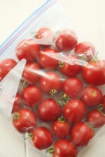 A gallon ziploc bag full of tomatoes sits on a white wooden table. The tomatoes still have stems on and are a beautiful ripe red. The bag is laying flat, ready to go to into the freezer.