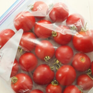 A gallon ziploc bag full of tomatoes sits on a white wooden table. The tomatoes still have stems on and are a beautiful ripe red. The bag is laying flat, ready to go to into the freezer.