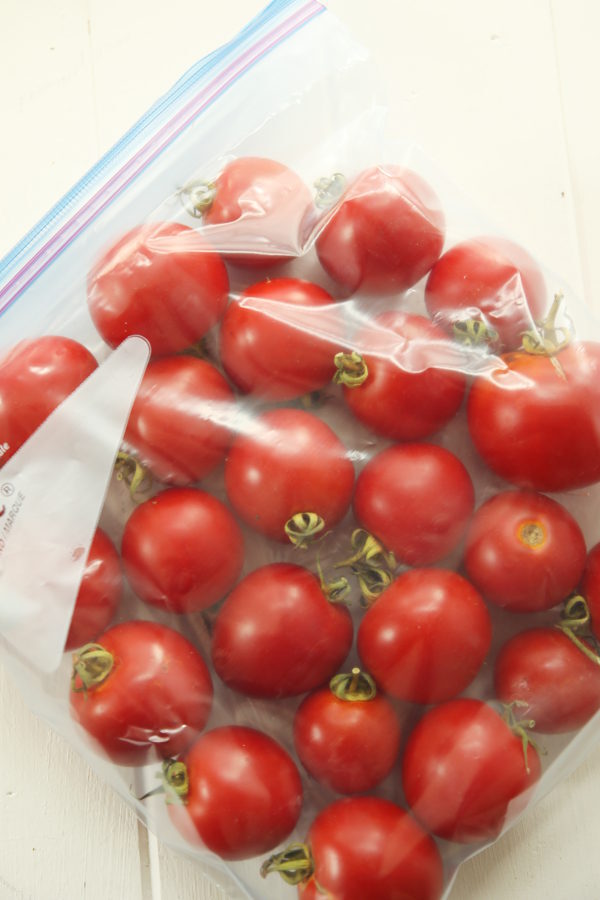 A gallon ziploc bag full of tomatoes sits on a white wooden table. The tomatoes still have stems on and are a beautiful ripe red. The bag is laying flat, ready to go to into the freezer.