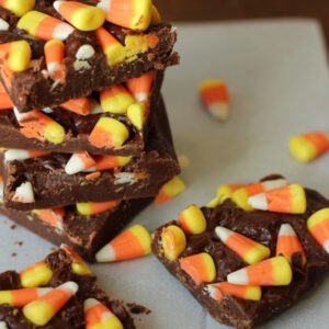 Squares of easy chocolate fudge are covered with candy corn. There is a stack of the candy corn fudge in the back with two squares of it sitting in the foreground. Candy corn is scattered around the setting.