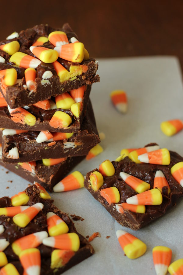 Squares of easy chocolate fudge are covered with candy corn. There is a stack of the candy corn fudge in the back with two squares of it sitting in the foreground. Candy corn is scattered around the setting.