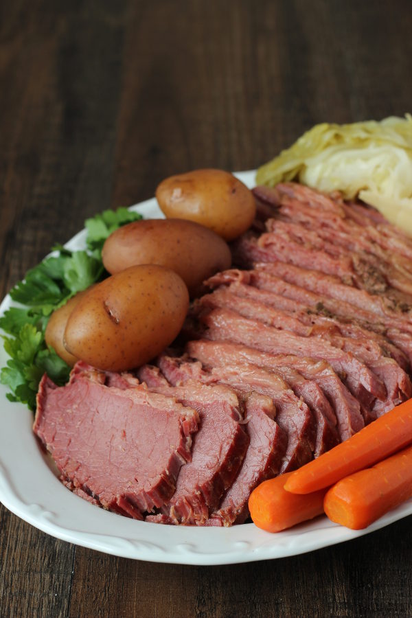 A wooden table holds a large white platter covered in corned beef dinner. There is a large corned beef sliced up, some small red potatoes, a few carrots and wedges of cabbage. The plate is garnished with parsely.
