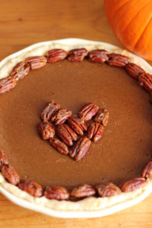 A homemade pumpkin pie sits on a wooden table in a white pie pan. The pie is garnished with candied pecans around the edge and in a heart shape in the middle. There is a pie pumpkin in the background of the photo.