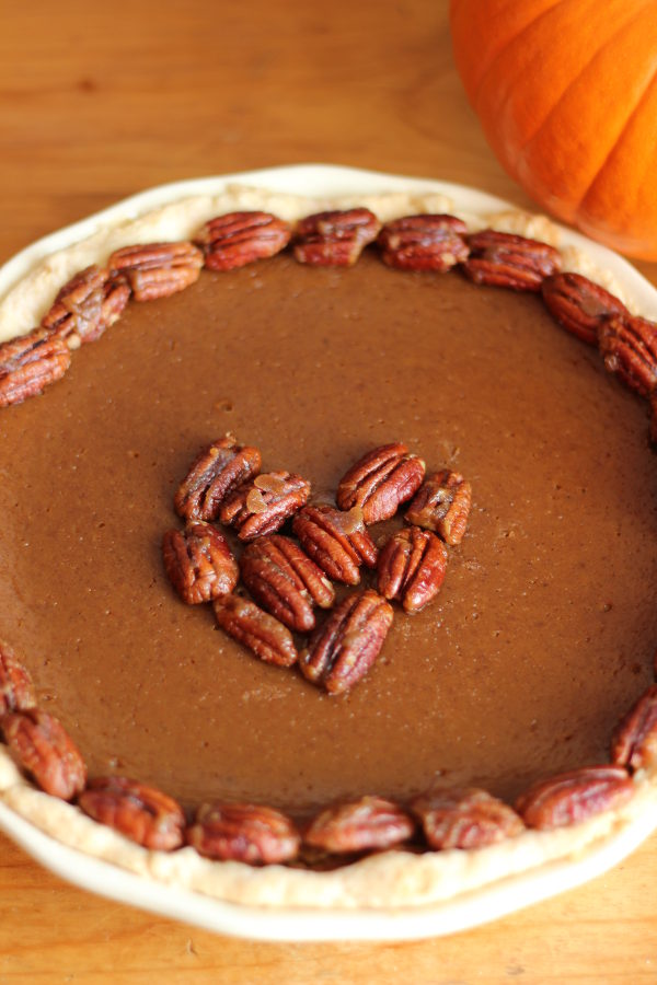 A homemade pumpkin pie sits on a wooden table in a white pie pan. The pie is garnished with candied pecans around the edge and in a heart shape in the middle. There is a pie pumpkin in the background of the photo.