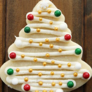 A cut out cookie in a Christmas tree shape is decorated with white buttercream and green, red, white and gold balls like ornaments.