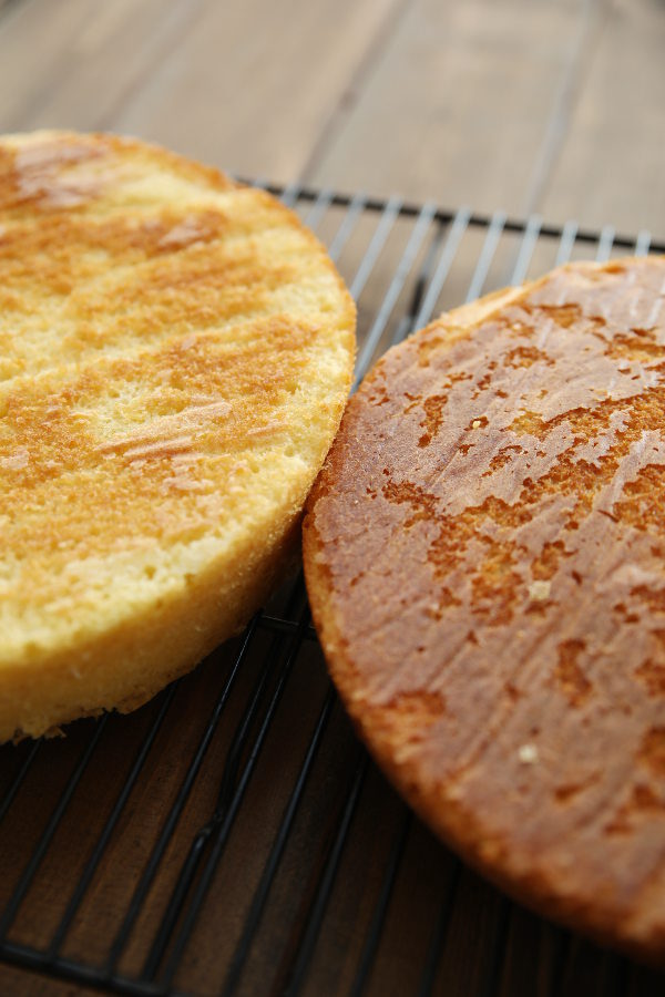 Two cakes are pictured upside down on a rack. The cake on the left is lighter in color because it was baked with cake strips compared to the cake on the right which is darker.