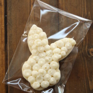 A bunny face and ears cut out sugar cookie is decorated with swirls of white frosting. It is packaged in a self sealing plastic bag for gifting.