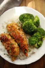 Two pieces of honey garlic chicken sit on a bed of rice next to a side of broccoli. The plate of chicken dinner is on a wooden table.
