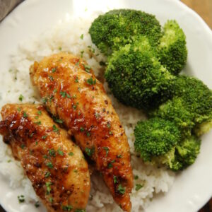 Two pieces of honey garlic chicken sit on a bed of rice next to a side of broccoli. The plate of chicken dinner is on a wooden table.