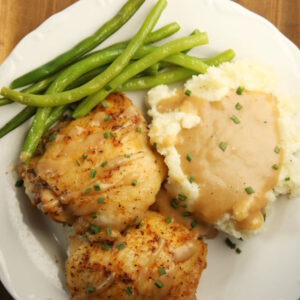 A white plate holds two golden, crispy baked chicken thighs, some green beans, and a pile of mashed potatoes with gravy.