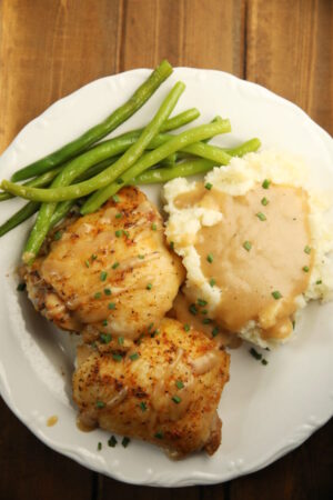 A white plate holds two golden, crispy baked chicken thighs, some green beans, and a pile of mashed potatoes with gravy.