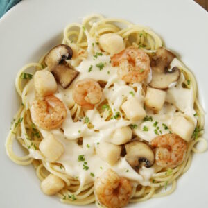 A white plate is full of tender shrimp, sweet bay scallops, and plenty of alfredo sauce drizzled over pasta.