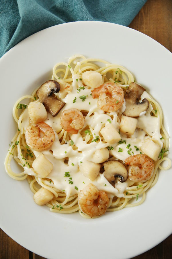 A white plate is full of tender shrimp, sweet bay scallops, and plenty of alfredo sauce drizzled over pasta.