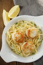 A round white dish holds buttery pasta topped with three large golden crusted scallops. There are wedges of lemon and a gray napkin nearby.