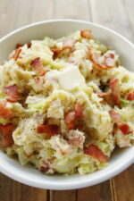 A large white bowl full of Irish colcannon. You can see large pieces of bacon and chunks of tender cabbage and a pat of butter melting on top.