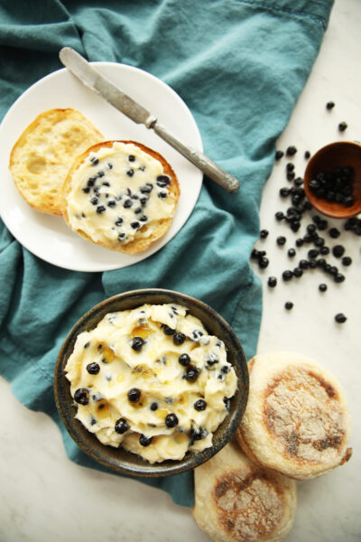 This is a photo with a potter bowl full of blueberry honey butter. This dried blueberry compound butter is also spread on some English Muffins and there is a small dish of dried wild blueberries. You can see a very light drizzle of honey on the top of the bowl of blueberry honey butter.