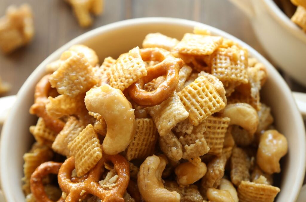 This is a bowl of chex mix snack mix on the table.  The mixture has lots of large cashews and some pretzels in with the chex.