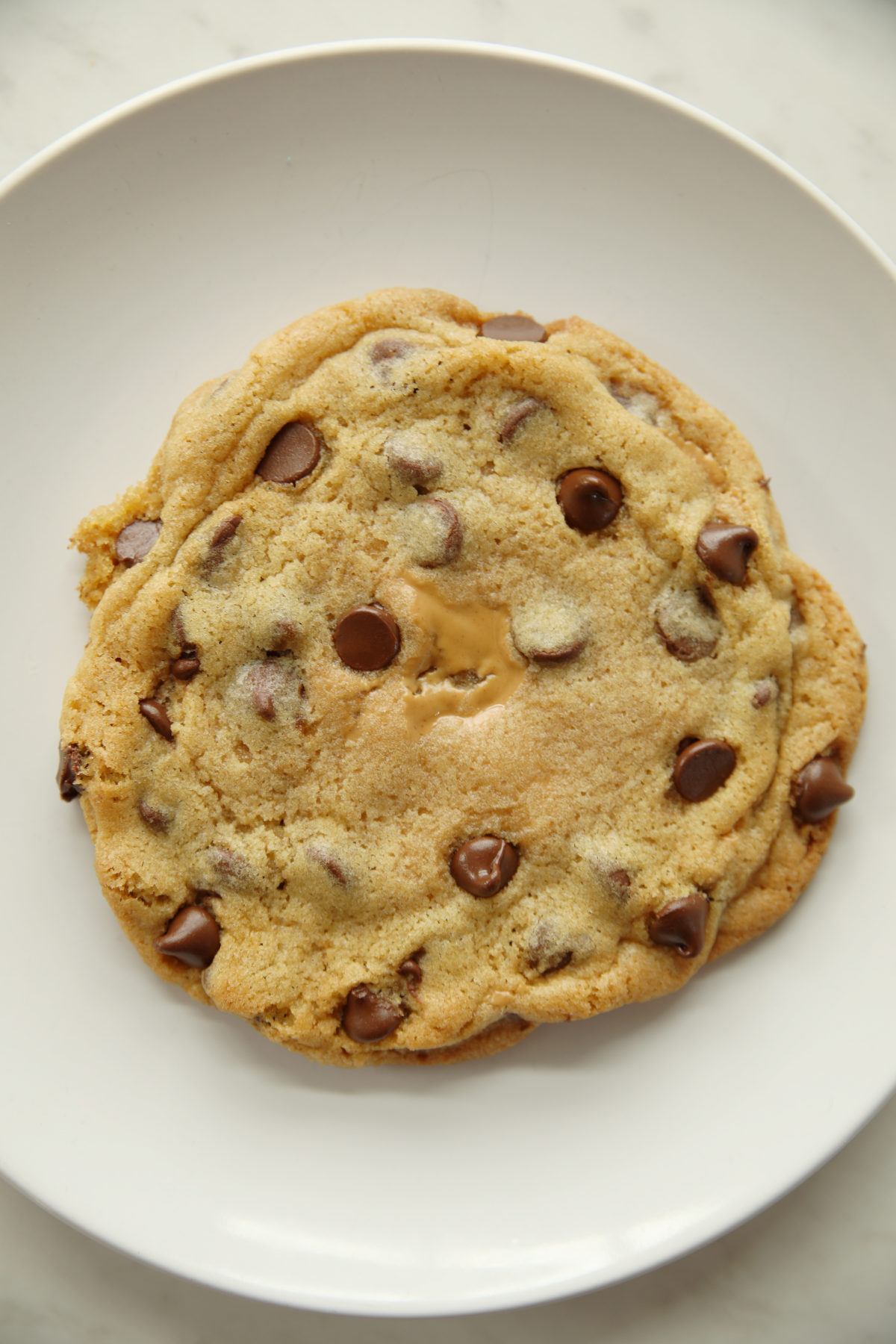 This is a photo of a large peanut butter stuffed chocolate chip cookie on a white plate. You can see the peanut butter peaking through the middle of the chocolate chip cookie.