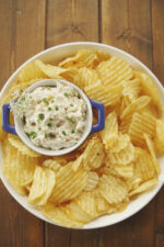 A big white dish is full of thick, crinkle cut potato chips. Nestled into the chips is a blue dish with little handles and it is full of creamy French onion dip. The dip is garnished with a chive flower and fresh chives.