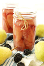 Two jars of pears canned in blackberry simple syrup. The jars are sitting on a cloth with some blackberries and fresh pears.