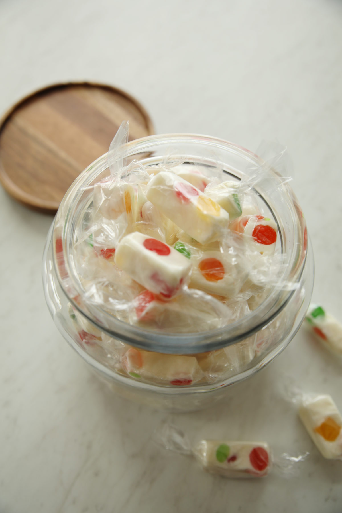 A pretty glass jar full of individually wrapped pieces of gumdrop jelly nougat. The jar has a wooden lid off to the side.