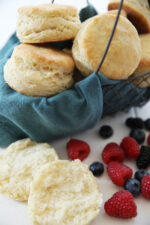 a beautiful basket lined with a dark blue cloth is full of flakey homemade biscuits. You can see a biscuit on the table open to the flakey layers and some fresh berries next to the biscuits.