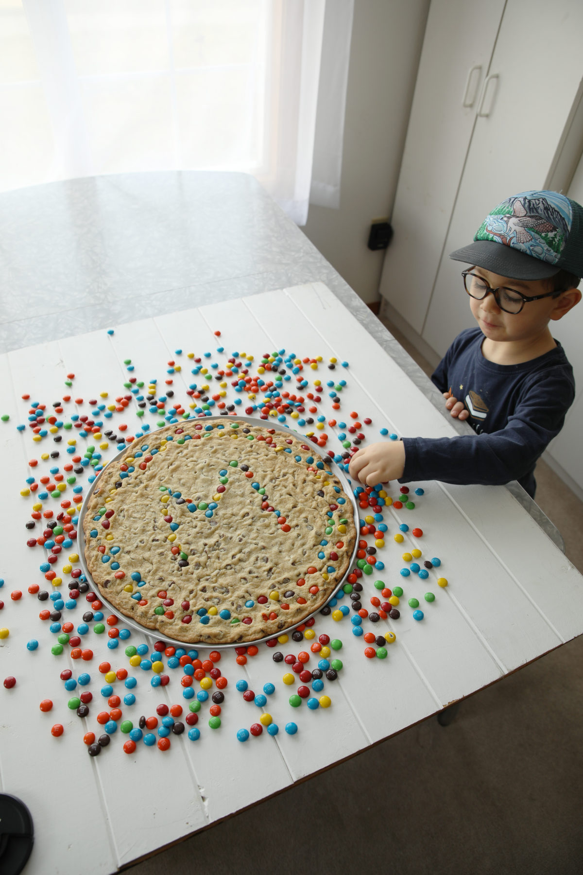 a photo of a photography studio where a 3 year old boy is grabbing an M and M off a table that also has a chocolate chip cookie cake on it. 