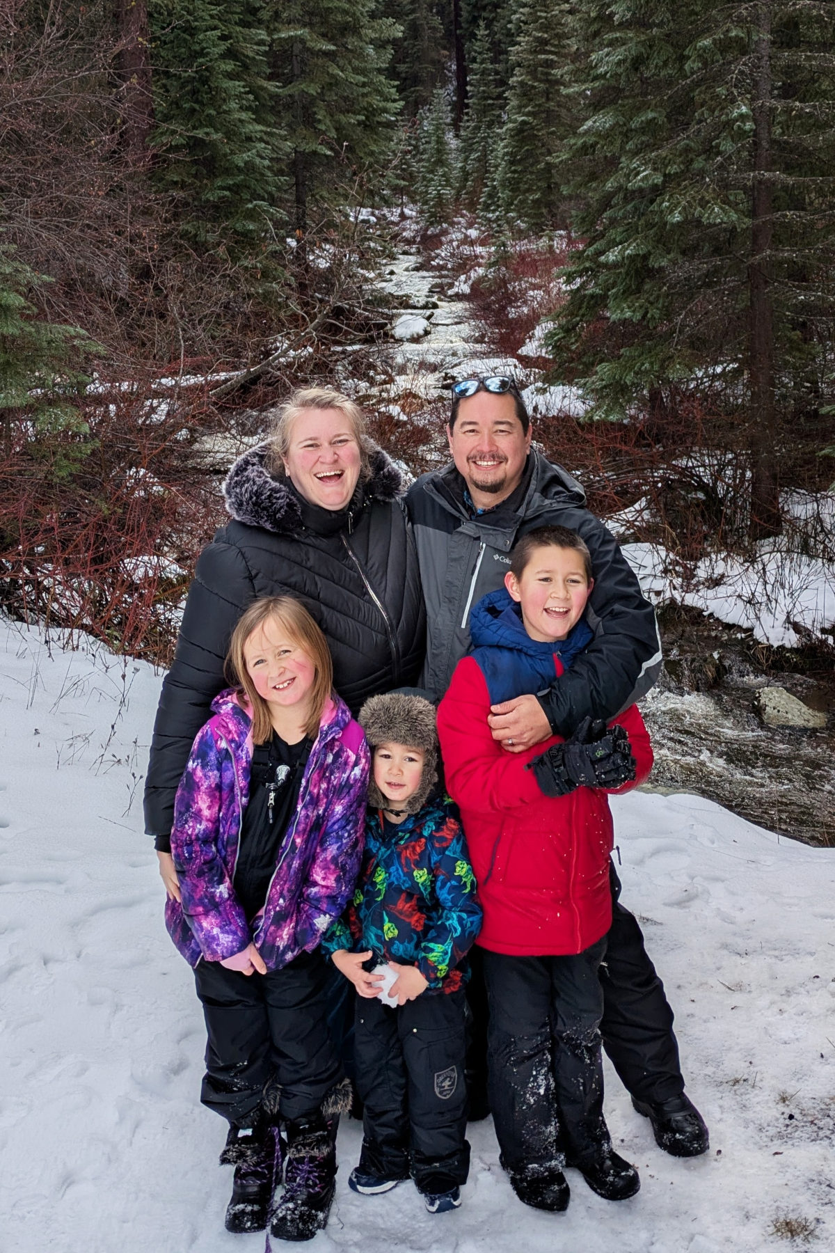 Mirlandra Neuneker with husband and three children in the snow in front of evergreen trees in the mountains. Everybody is bundled up in coats for the winter.