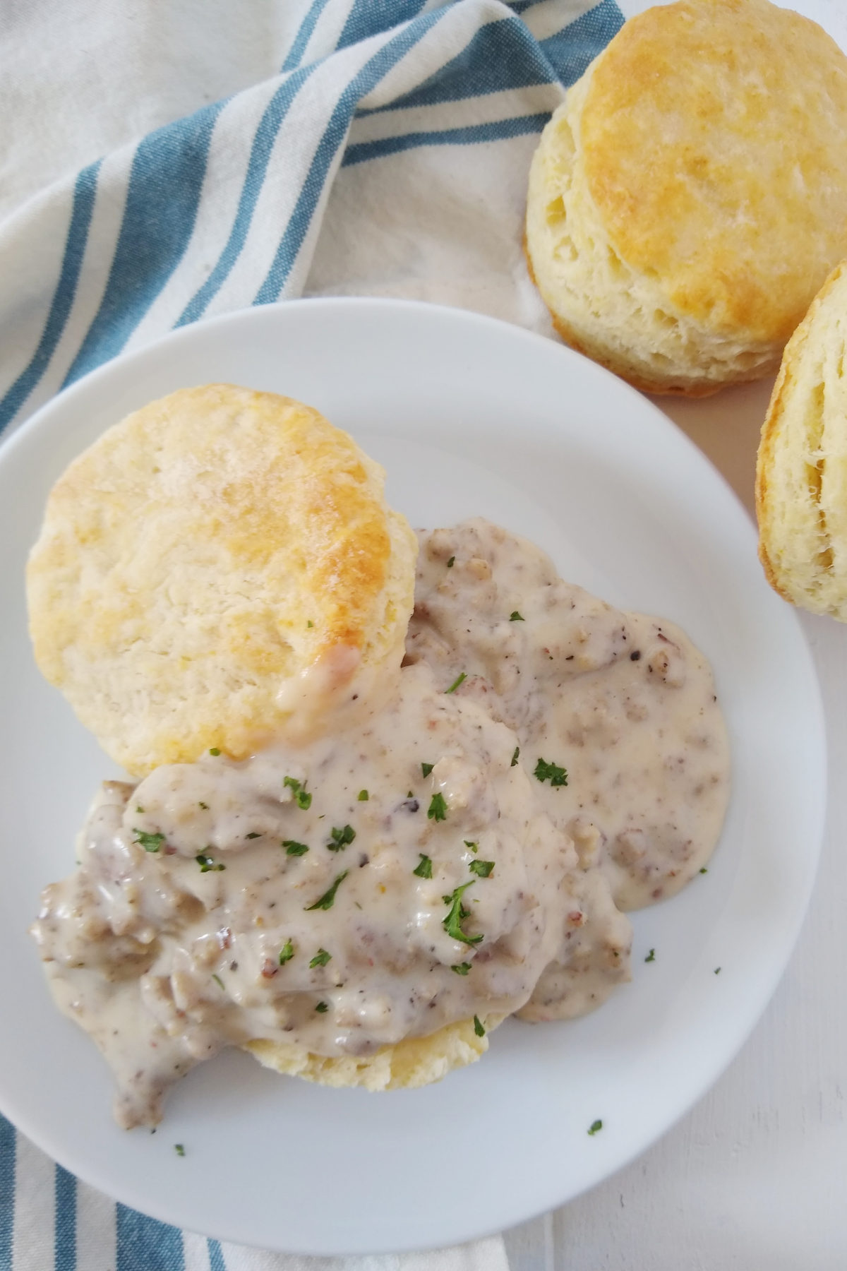 A picture of my amazing biscuits and gravy recipe. You can see a split biscuit slathered in homemade sausage gravy. There are a few green herbs on top as garnish and more biscuits on the side.
