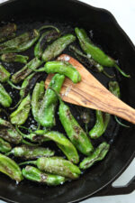 A cast iron skillet full of blistered shishito peppers and an olive wood spatula.