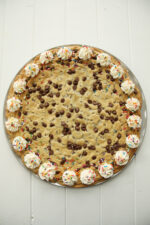 This beautiful chocolate chip cookie cake is baked in a 14" pizza pan and studded with extra chocolate chips on top. There is a whipped cream border with festive sprinkles on it.